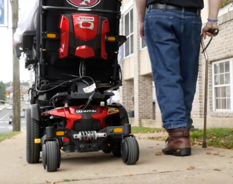 Person with cane walks with person in motorized wheelchair.