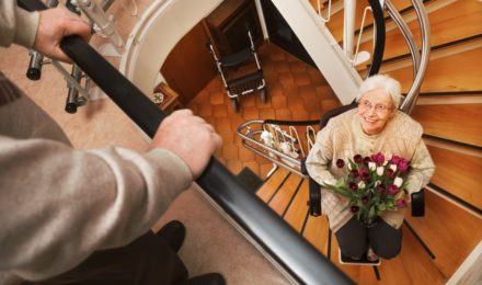 Elderly woman smiling looking up while holding a vase of flowers in a stair lift.